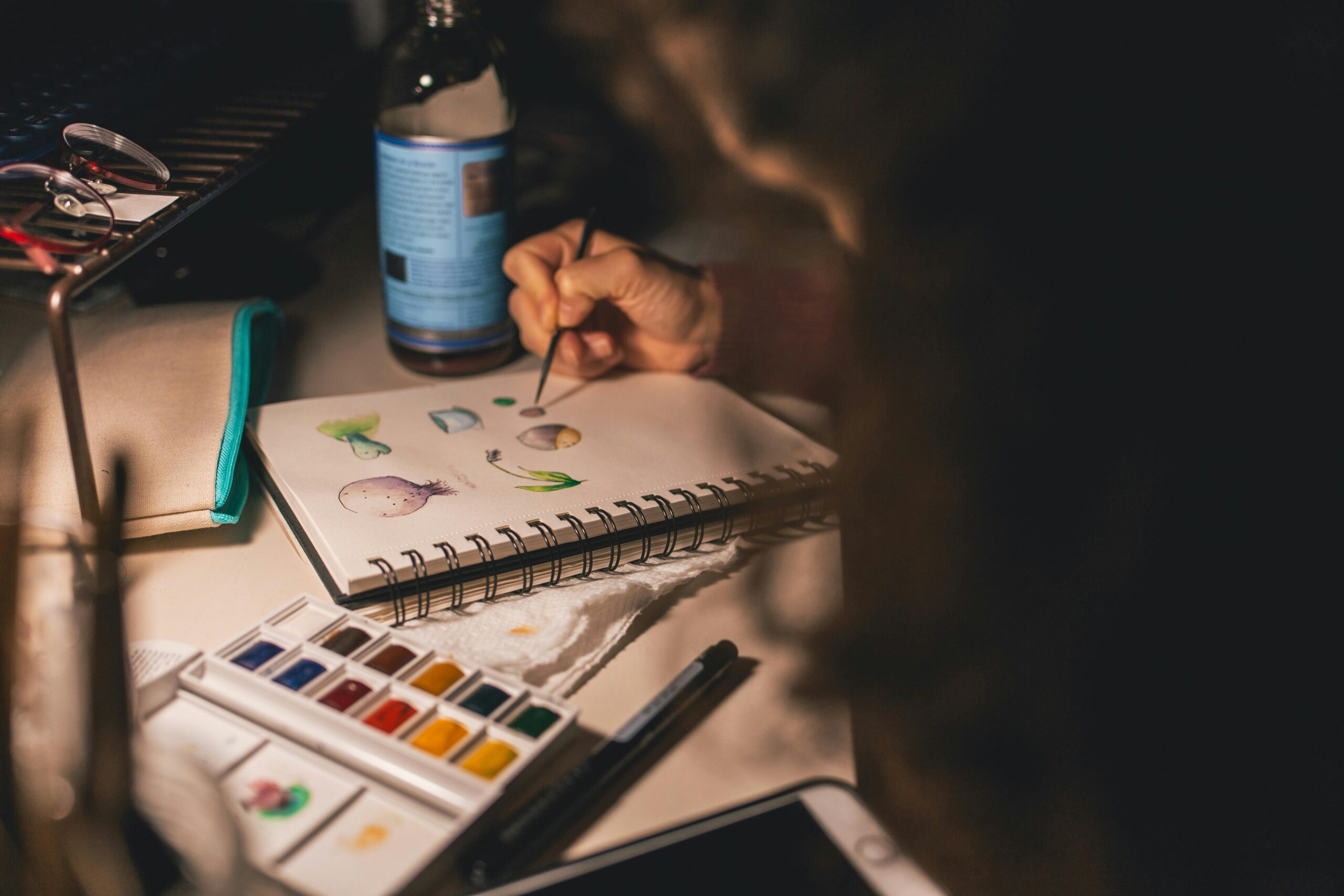 Artist painting with watercolors in a notepad at a desk. Creative process indoors.