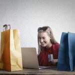 A joyful woman shopping online using a laptop with colorful shopping bags surrounding her.