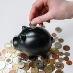 Hand inserting coin into a black piggy bank surrounded by various coins, symbolizing savings.