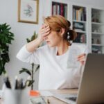 Worried woman counting expenses on a laptop while holding a receipt at her desk.